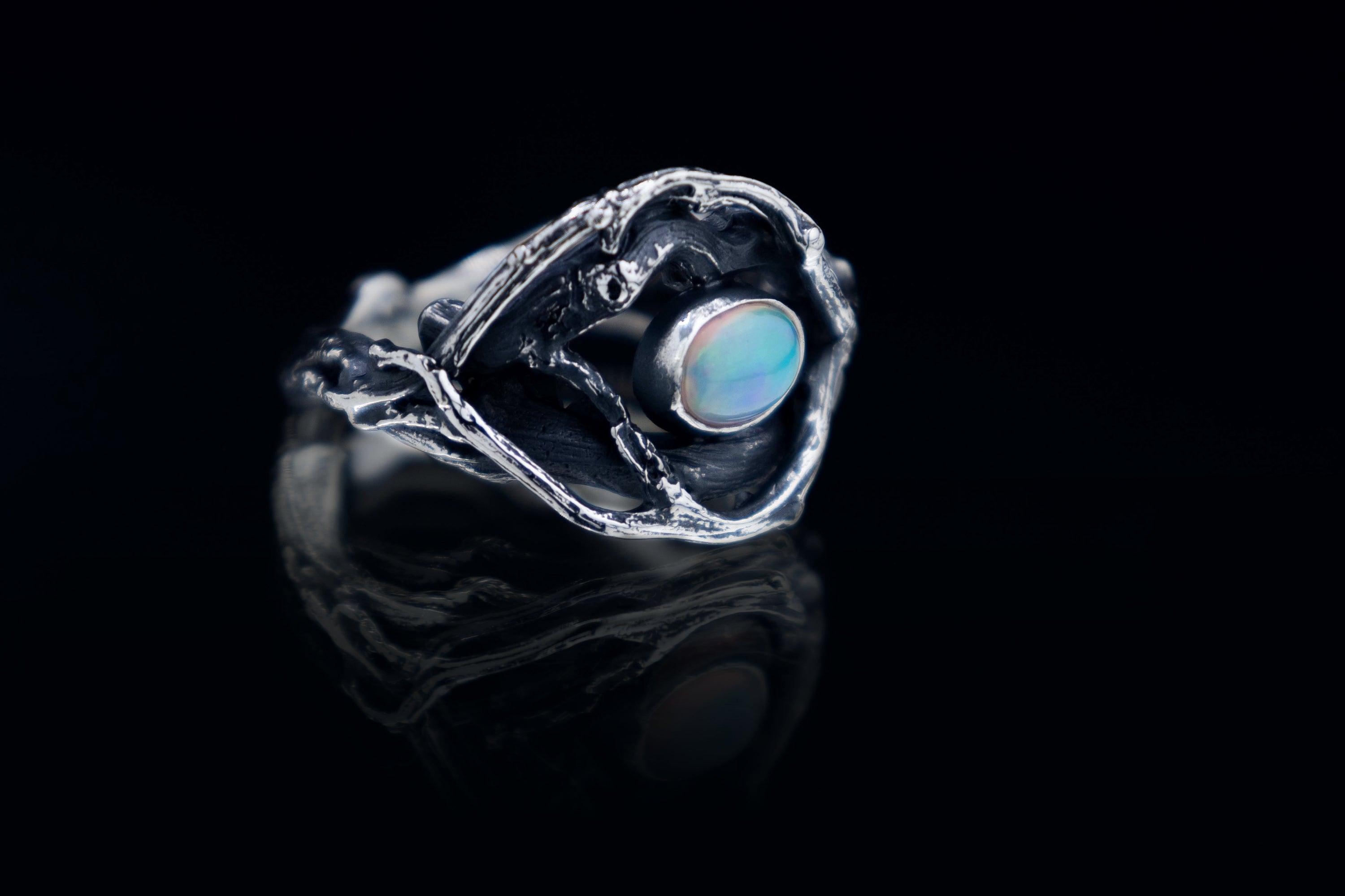 A silver ring with an oval, iridescent gemstone set within a textured, organic-looking frame. The ring's design resembles a stylized flower or open form, with intricate details and a polished finish. The gemstone has a blue and green color.