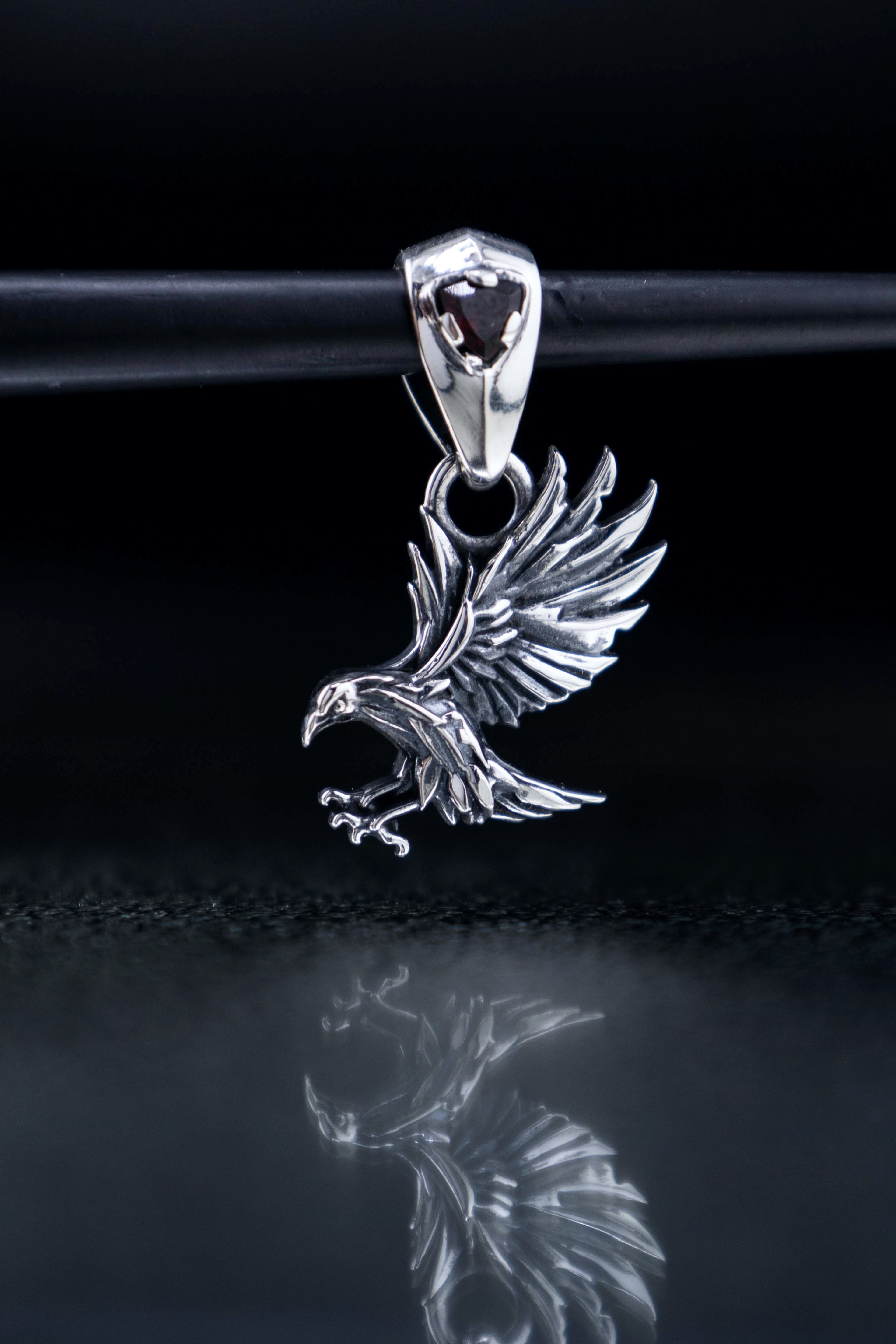 Sterling silver raven pendants on black reflective glass, gothic crow necklaces with blue or red gemstone bails, mythical bird jewelry artifact.