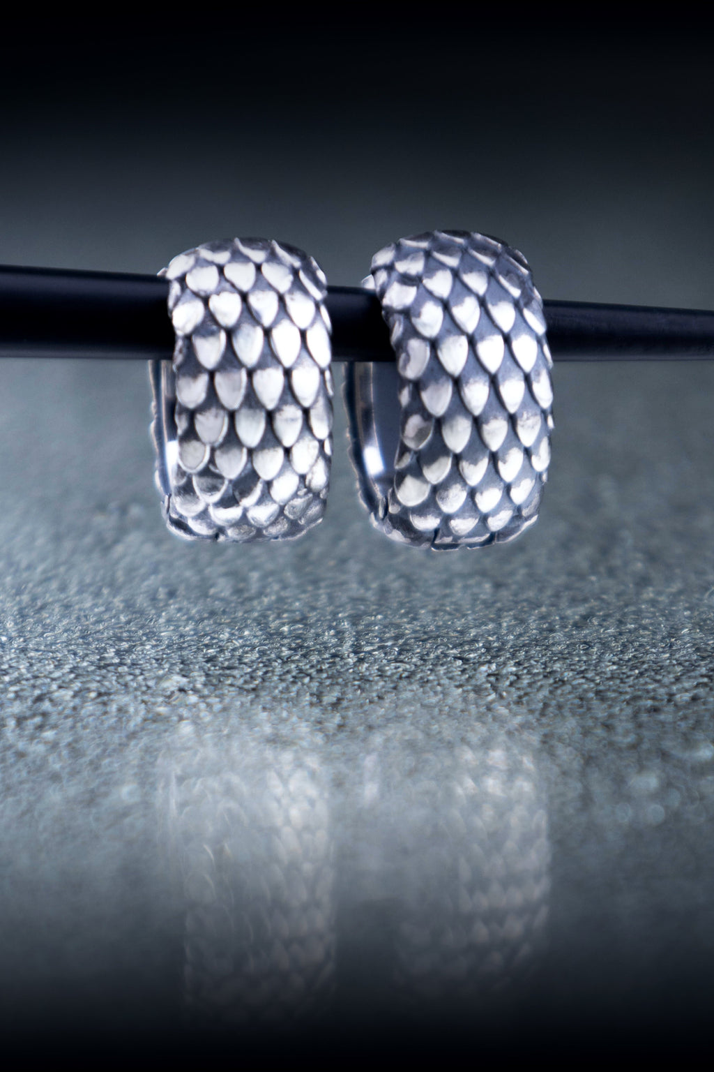 Two wide sterling silver hoop earrings with a repeating scale pattern hanging from a thin black rod over a reflective gray surface.