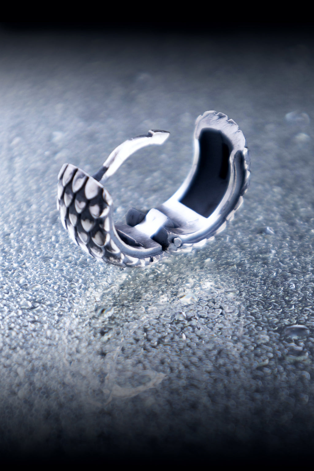 A single open silver hoop earring with a dragon scale pattern, showing the secure hinge and clasp mechanism on a reflective surface.