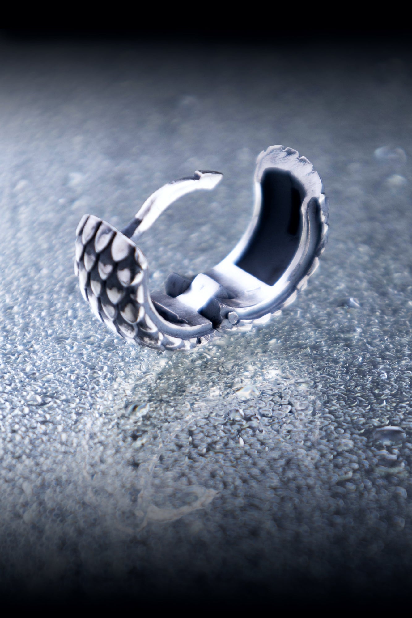 A single open silver hoop earring with a dragon scale pattern, showing the secure hinge and clasp mechanism on a reflective surface.