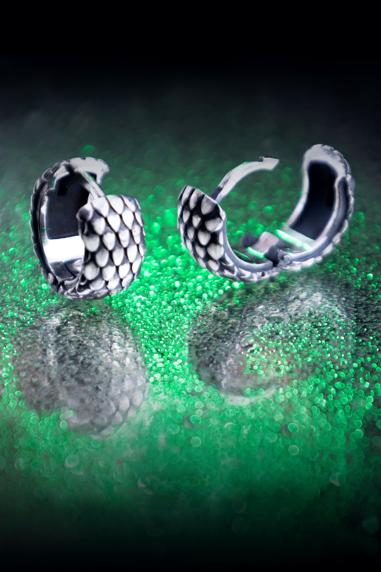 A pair of silver hoop earrings with a detailed dragon scale pattern, resting on a dark surface with emerald green bokeh and reflections.