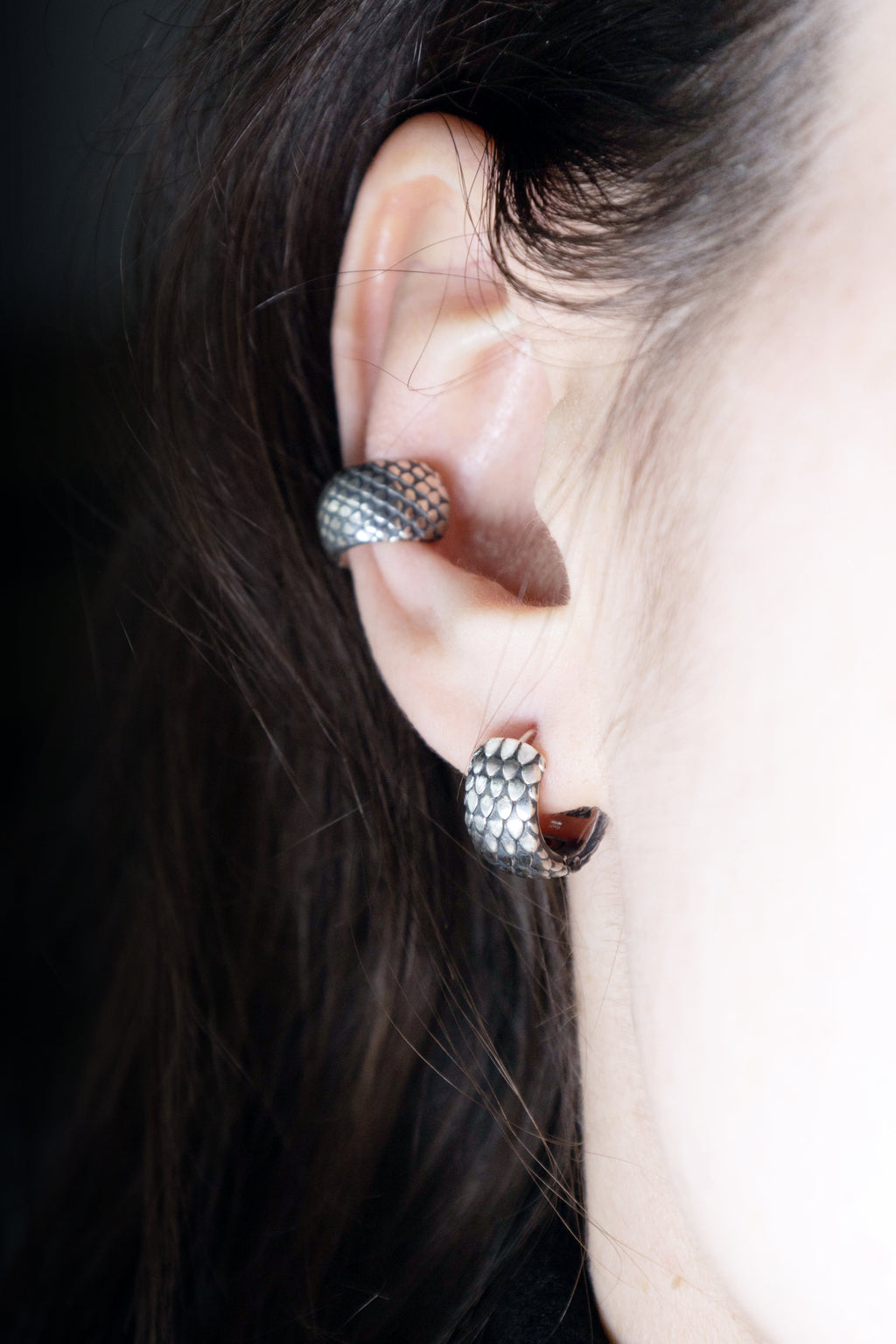 Close-up of a person's ear featuring two silver pieces: a small dragon scale hoop in the lobe and a textured scale ear cuff on the cartilage, framed by dark hair.