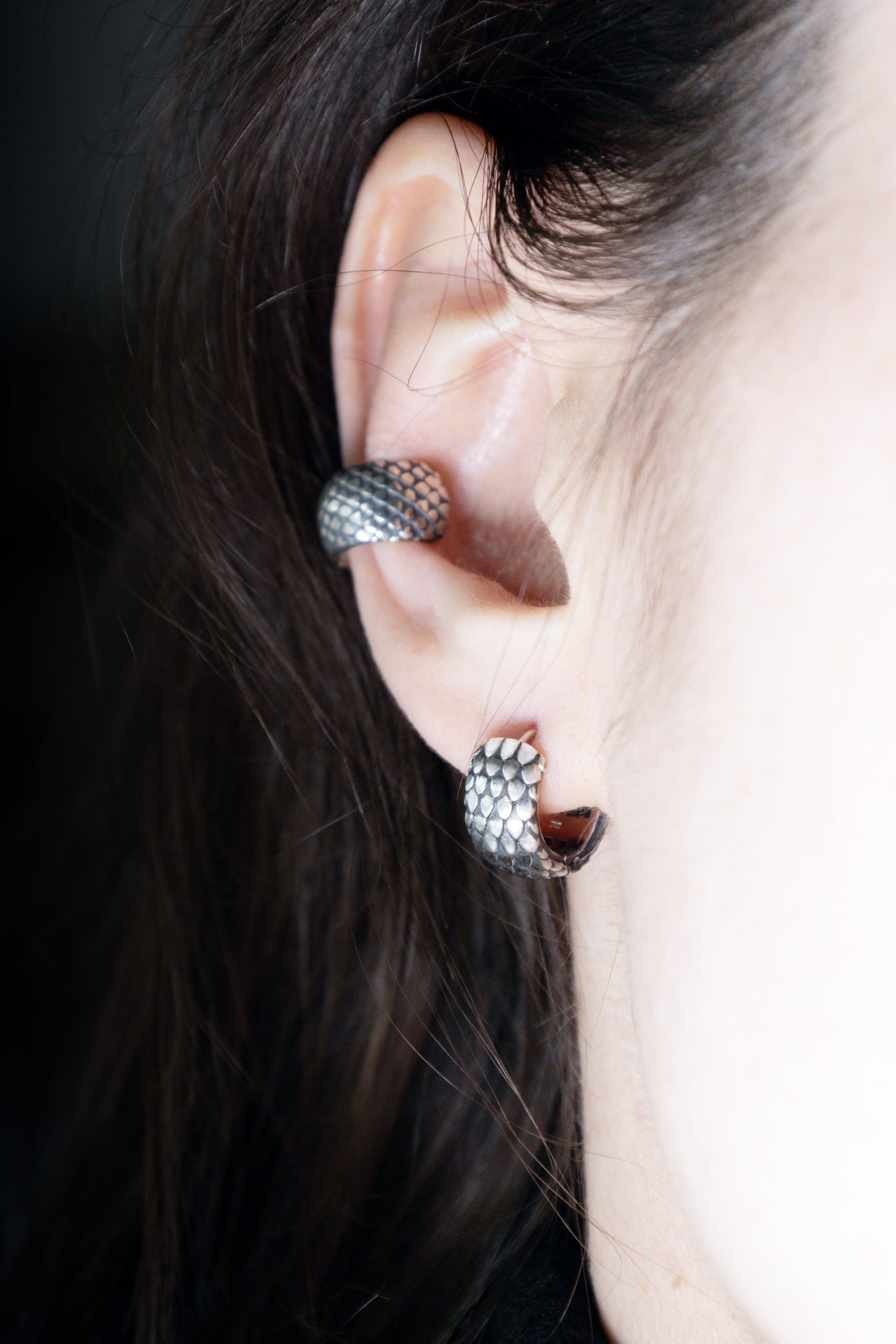 Close-up of a person's ear featuring two silver pieces: a small dragon scale hoop in the lobe and a textured scale ear cuff on the cartilage, framed by dark hair.