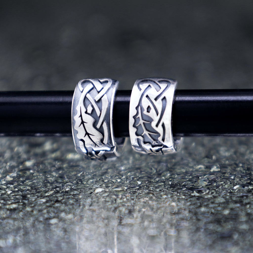 Artisan sterling silver oak leaf hoop earrings hanging on a display rod, showcasing detailed Celtic knots and mirrored reflections on a wet textured surface.