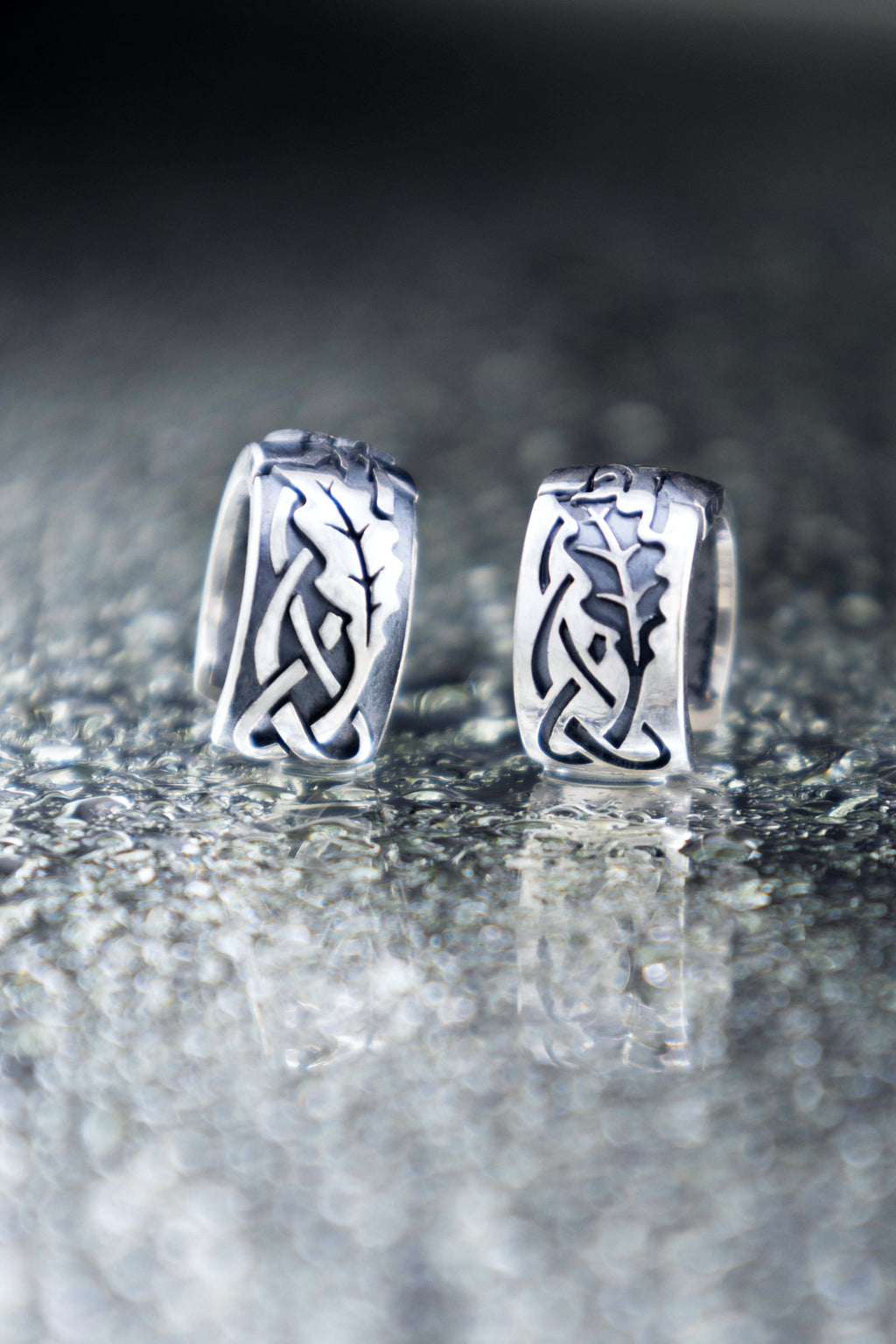 Close up of mismatched silver oak leaf huggie earrings on a glossy wet surface with bokeh light circles, highlighting the contrast of raised and engraved details.