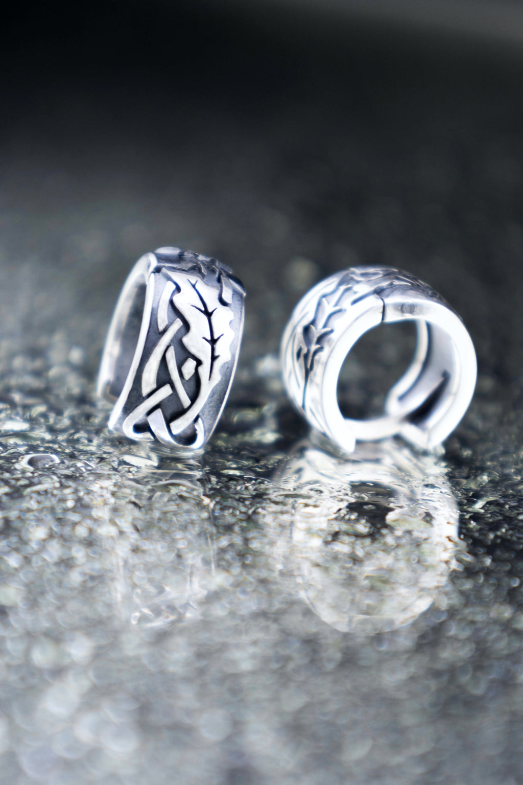 Close up of mismatched silver oak leaf huggie earrings on a glossy wet surface with bokeh light circles, highlighting the contrast of raised and engraved details.