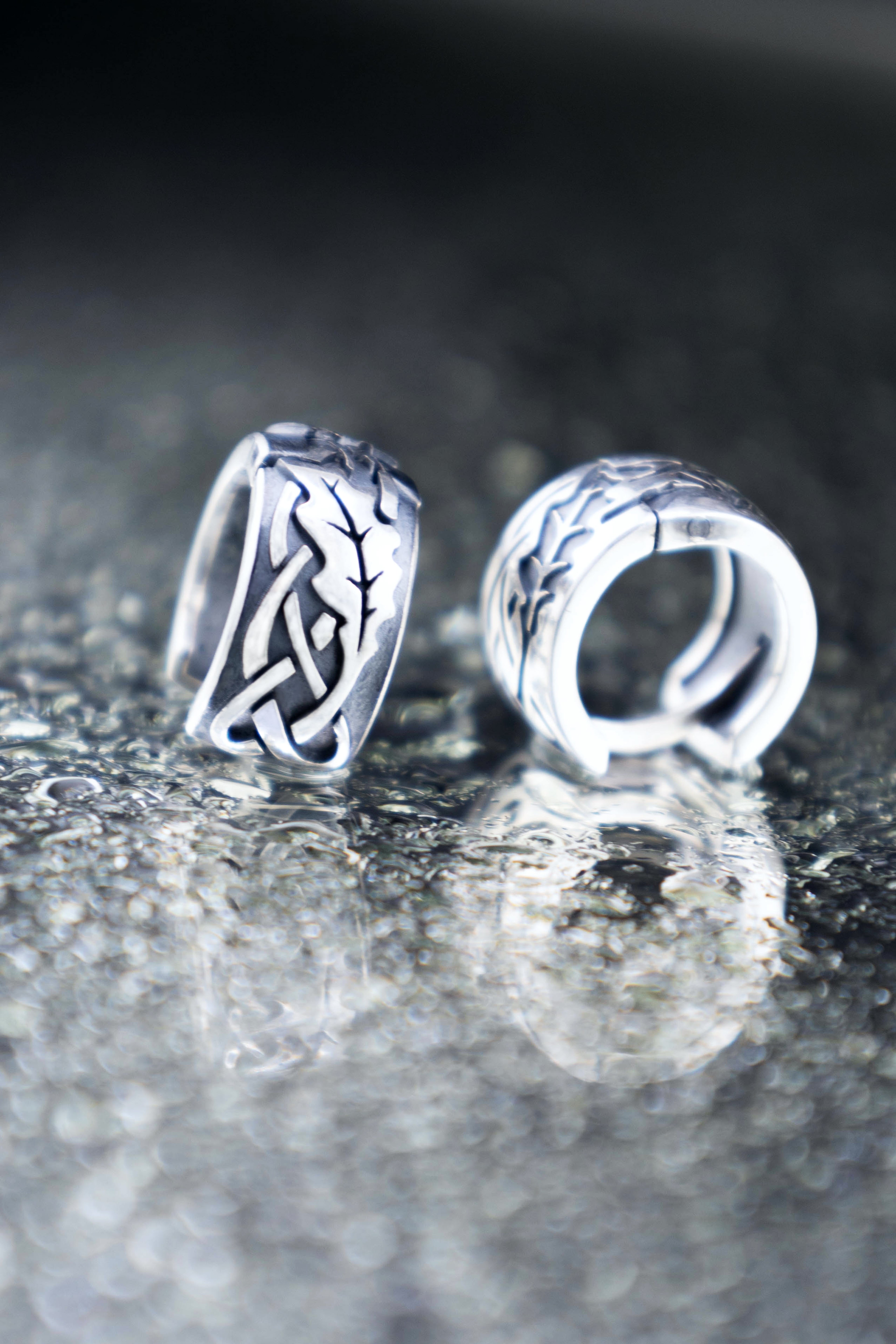 Close up of mismatched silver oak leaf huggie earrings on a glossy wet surface with bokeh light circles, highlighting the contrast of raised and engraved details.