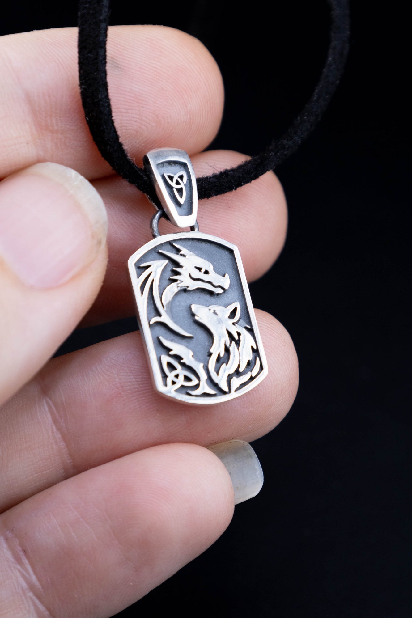 Sterling silver dragon and wolf pendant hanging on black cord against black background, full detail view of raised design and oxidized finish
