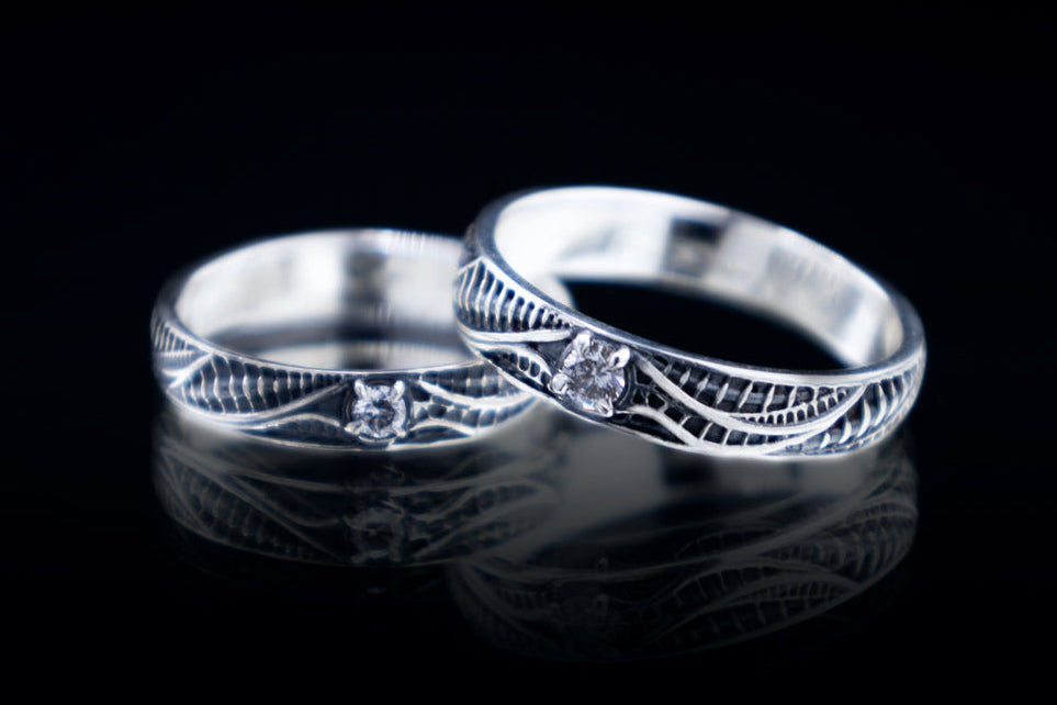 Two biomechanical sterling silver rings on a black reflective surface, each with a round lab-grown diamond and polished lines over dark ribbed recesses.