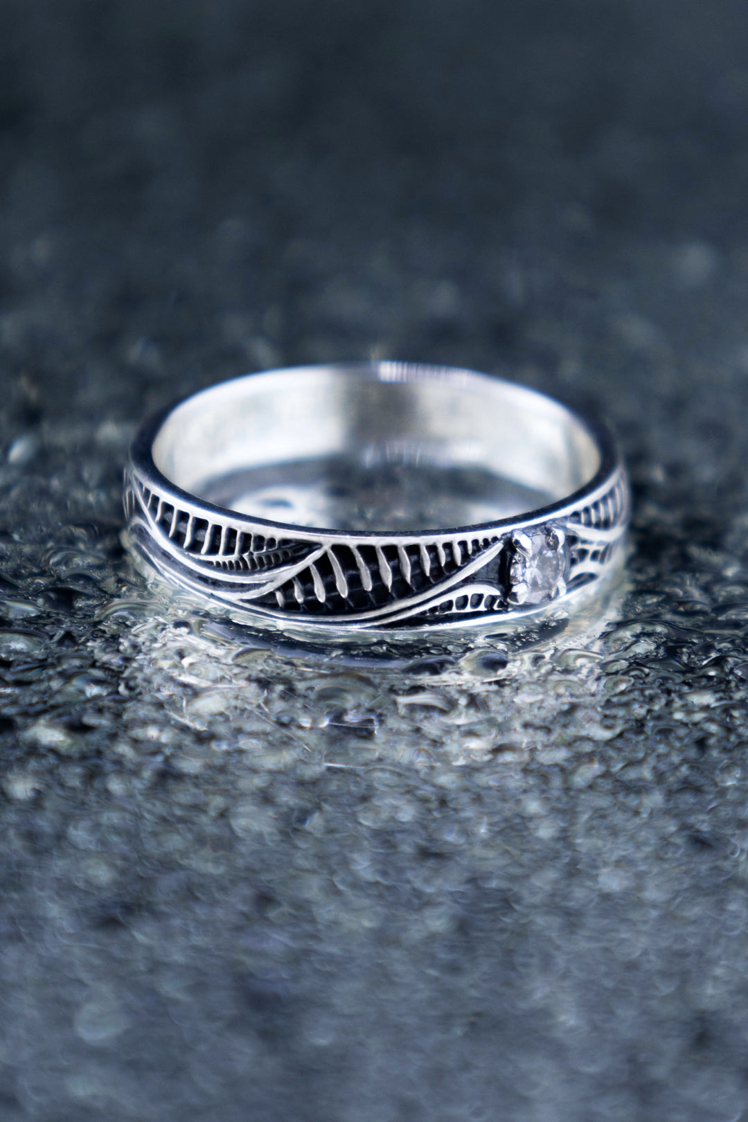 Angled close-up of a biomechanical silver ring with flowing polished lines, dark ribbed recesses and a round lab-grown diamond on a textured reflective surface.