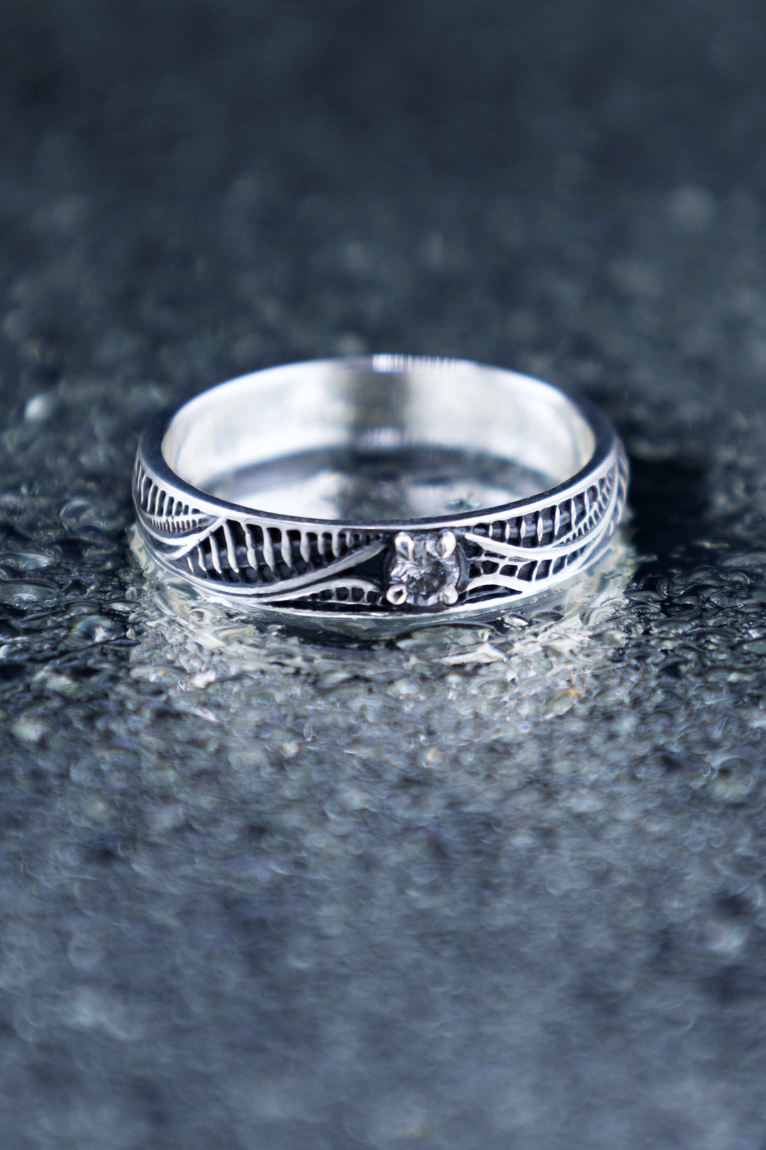 Front view of a biomechanical sterling silver ring on a textured reflective surface, with a round lab-grown diamond framed by flowing silver lines.